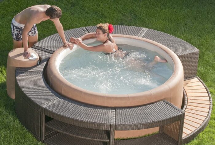 Heller runder Whirlpool mit Polyrattan Umrandung und Holztreppeeinstieg. Eine Frau badet und ein Mann steht am Rand des Whirlpools.