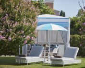 Der Unosider Pavillon Venice Cabana in einem Garten mit zwei Liegen und einem blau-weiss gestreiften Sonnenschirm.