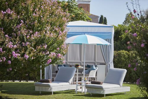 Der Unosider Pavillon Venice Cabana in einem Garten mit zwei Liegen und einem blau-weiss gestreiften Sonnenschirm.