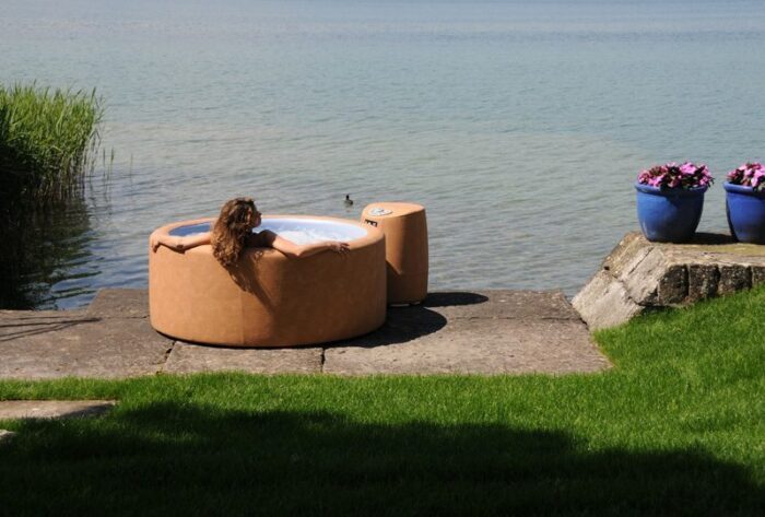A woman bathes in a round Softub Sportster model in a brown colour. The Softub is in front of a lake.