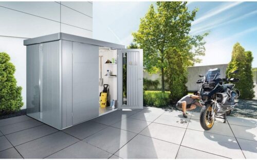 A silver metallic garden shed in front of a big house standing on a stony floor. In front of the garden shed is a man with his motorcycle and in the garden you can see different trees.