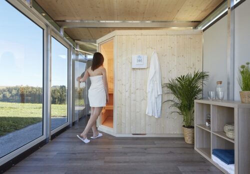 A woman wrapped in a white towel is entering the sauna through a corner entry. The sauna module is built into the outbuilding CasaNova.