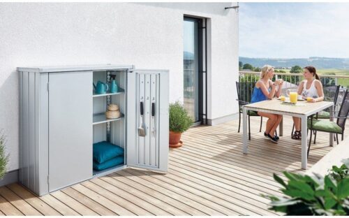 Silver patio locker with open door. Terrace storage box standing on a wooden terrace in front of a big, white house.