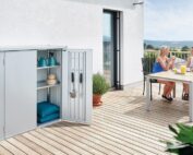 Silberner Terrassenschrank mit offen, stehender Tür. Terrassenschrank steht auf einem Holzbalkon vor einer weissen Wand.