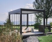 Dunkelgraue, freistehende Pergola mit verstellbaren Dach- und Seitenlamellen in einem grossen Garten.