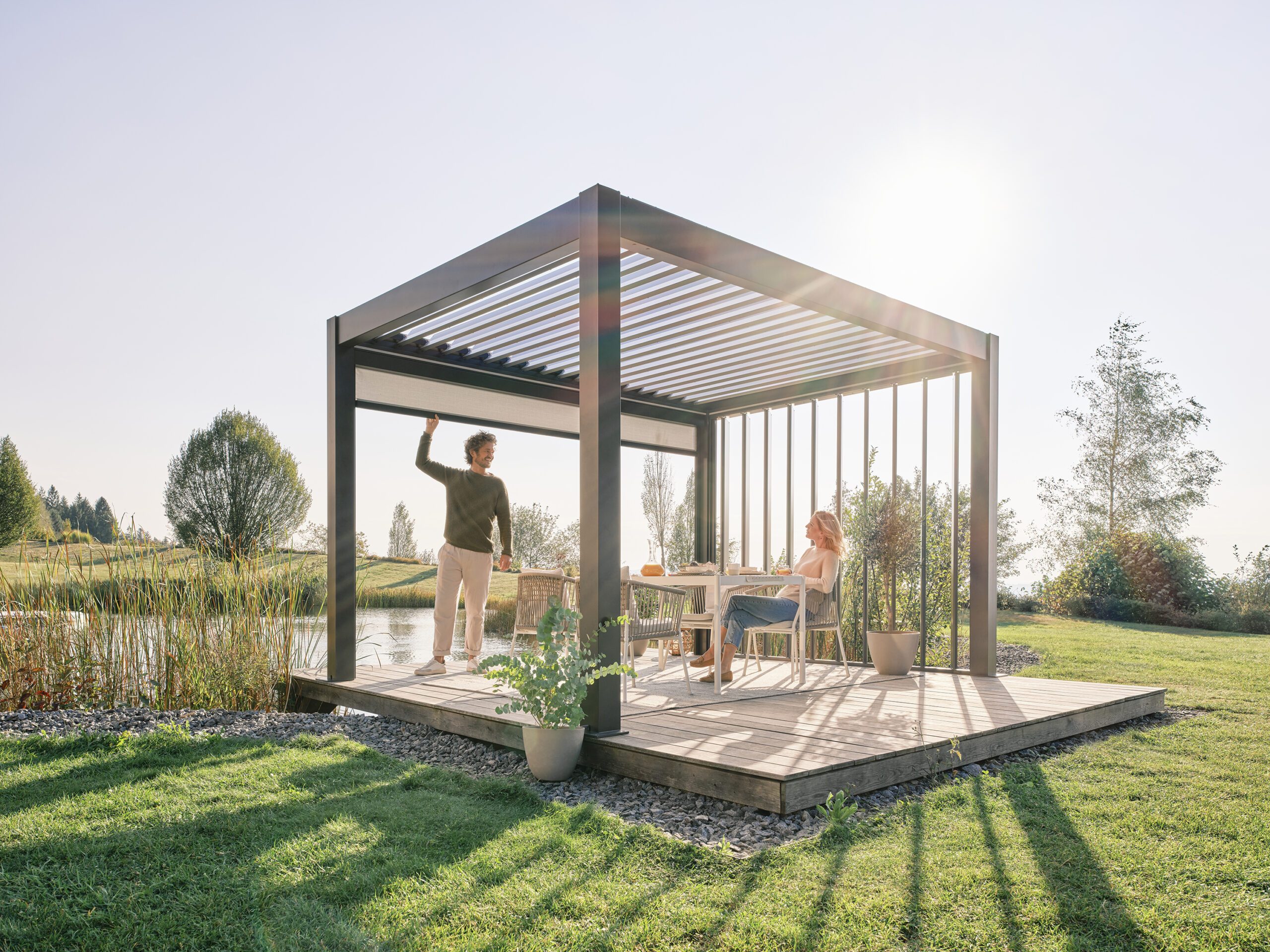 Pergola indépendante gris quartz dans un jardin avec un store solaire et des lamelles de toit ouvertes. Sous la pergola se trouve une table de jardin blanche à laquelle est assise une femme et un homme qui tire le store vers le bas.