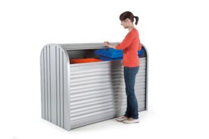 Biohort_StoreMax_160_silber_oeffnen_Muelltonne_Anwendungsbeispiel_Softub_Schweiz Frau steht vor einer silbernen Gartenbox mit rotem und blauem Container und öffnet den Rollladen