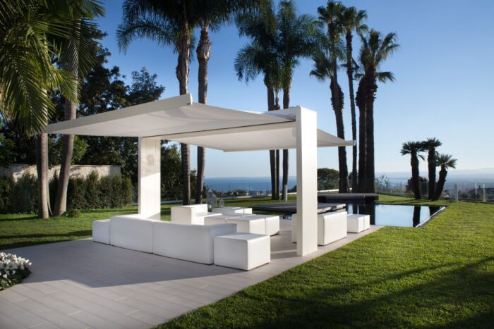 Unosider Pavillon Gate Shade weiss. Perspektive von unter dem Dach mit Gartensitzplatz im Schatten mit Blick auf Pool, Meer und Palmen.