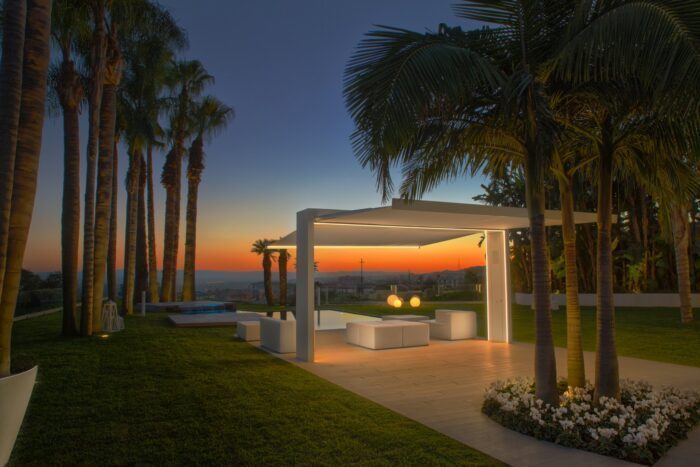 Weisser Unosider Gate Shade Pavillon im dunkeln bei Sonnenuntergang mit Palmen im Garten auf der Terrasse.