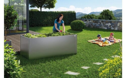 A grey raised vegetable bed, standing on a lawn. A woman is doing the gardening and two kids are playing in the background.
