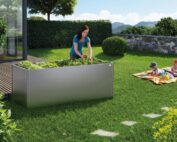 Ein graues Biohort Pflanzenbeet. Pflanzenbeet steht auf einem Rasen. Eine Frau gärtnert, während zwei Kinder auf einer Decke im Garten spielen