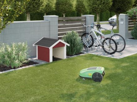 Weka Quality Mower Garage . A small garden with a green robotic lawn mower and a little red house that serves as a garage for the robot. Next to the little house are two bicycles and a white fence behind it.
