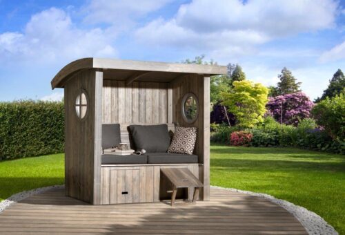 a wooden made seat arbour with grey cushions and wooden drawers underneath. The seat arbour stands in a garden and has two rounded windows on each side.