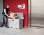 Postbote legt Pakete in silberne Gartenbox vor einem Haus