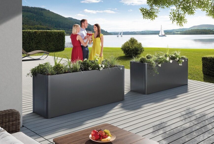 Abgebildet sind zwei dunkelgraue metall Pflanzenbeete von Biohort. Drei Personen stehen vor den Pflanzenbeeten. Pflanzenbeete stehen auf einer Terrasse vor einem See.