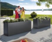 Zwei dunkelgraue Metall Pflanzenbeete von Biohort. Drei Personen stehen vor den Pflanzbeeten. Pflanzbeete stehen auf einer Terrasse vor einem See.