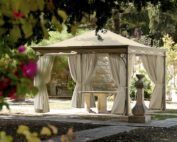 Der Unosider Pavillon Novecento Rain in einem grossen Garten mit spitzem Dach und zusammengebundenen Vorhängen vor einem Pool.
