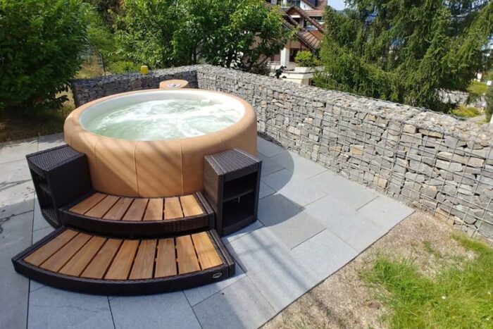 A brown round Sportster soft tub in a garden, with polyrattan and wooden staircase entrance.