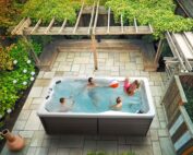 Eine Familie spielt mit einem Ball in einem Swim Spa mit grauer Aussen- und weisser Innenfarbe. Der Swim Spa steht in einem Garten