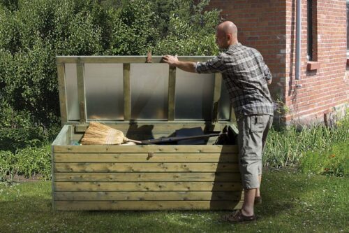 Ein Mann öffnet eine naturbelassene Terrassentruhe aus Holz. Terrassentruhe mit einem durchsichtigen Dach. Truhe steht in einem Garten.