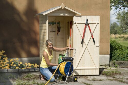 a woman is putting away the garden hose. She is kneeling in front of a narrow garden cabinet with a saddle roof and wooden natural look. The cabinet is filled with different garden tools.