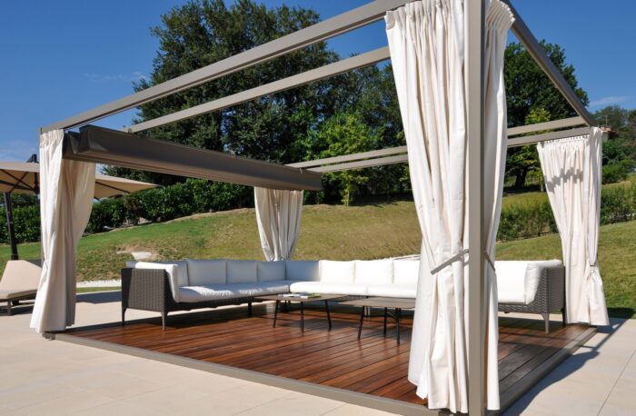 Unosider Pavillon Mood mit weissen, zusammengebundenen Vorhängen auf einer Terrasse mit Holzboden in einem Garten mit Pool.