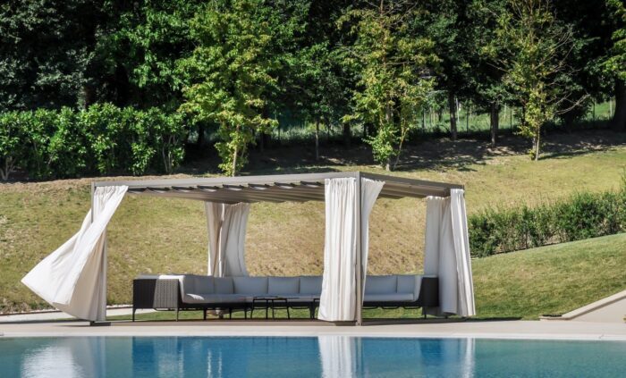 Unosider Pavillon Mood mit weissen, zusammengebundenen Vorhängen auf einer Terrasse mit Holzboden in einem Garten mit Pool.