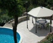 Der Unosider Pavillon Novecento Esagono steht neben einem Pool in einem prachtvollen Garten mit grossen Bäumen und Büschen. Der pavillon hat ein weisses spitz Dach und darunter stehen Stühle und ein Tisch aus Stahl in italienischem style.