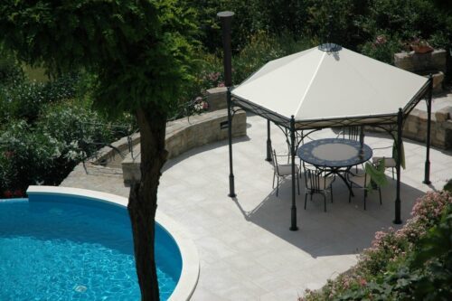 Le pavillon Novecento Esagono d'Unosider se trouve à côté d'une piscine dans un magnifique jardin avec de grands arbres et arbustes. Le pavillon a un toit blanc pointu et en dessous se trouvent des chaises et une table en acier de style italien.