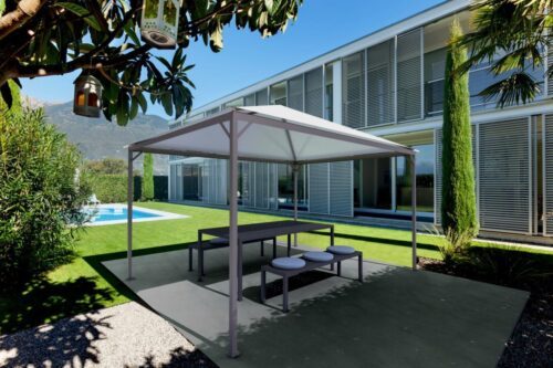 Pavillon avec des poutres fines et un toit blanc. Couvert d'une table et d'un banc. Le pavillon se trouve devant un bâtiment avec une pelouse et une piscine.