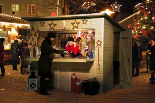 Ein verkaufshaus an einem Weihnachtsmarkt. EIne Frau mit einer roter Jacke steht im Verkaufshaus und bedient grade einen Kunden. Das Verkaufshaus ist beschmückt mit Sternen und Lichter.