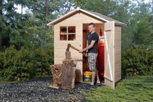 Kleines Gerätehaus in naturbelassener Farbe mit Fenster, Einzeltür und Satteldach. Steht auf einem Steinboden im Garten. Vor dem Haus schlägt ein junger Mann Holz.