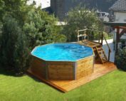 Weka Massivholzpool mit Sandfilteranlage. Ein Pool aus Massivholz steht in einem Garten auf einem Holz boden in der gleichen Farbe. Der Swimmingpool ist gefüllt und hat auch eine Holztreppe mit einer silbernen, metallen Leiter für den Einstieg ins Wasser. Pool steht vor einem weissen Haus.