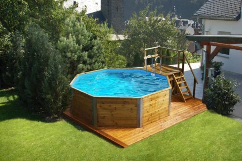 Weka solid wood pool with sand filter system. A pool made of solid wood stands in a garden on a wooden floor in the same colour. The pool is filled up and also has wooden steps with a silver metal ladder to get insider. The pool is in front of a white house.