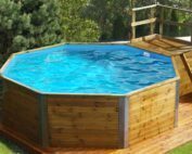 Weka Swimmingpool. Ein Swimmingpool aus Holz steht im garten vor einem weissen Haus auf einem Holzboden. Der Pool ist gefüllt mit Wasser und hat eine Holztreppe auf der rechten Seite für einen einfacheren Einstieg.