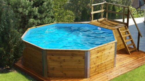 Weka Swimming Pool. A wooden swimming pool stands in the garden in front of a white house on a wooden floor. The pool is filled with water and has a wooden staircase on the right side for easier access.