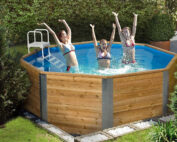 Ein Ein acht eckiger, hölzener Swimmingpool in einem Garten. Der Swimmingpool ist gefüllt mit Wasser und hat einen weisse Leiter als Übereinstieg. Drei Kinder stehen im Swimmingpool und haben Spass.