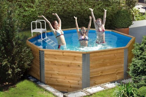 An eight-sided, wooden swimming pool in a garden. The swimming pool is filled with water and has a white ladder as an entrance. Three children are standing in the swimming pool and having fun.