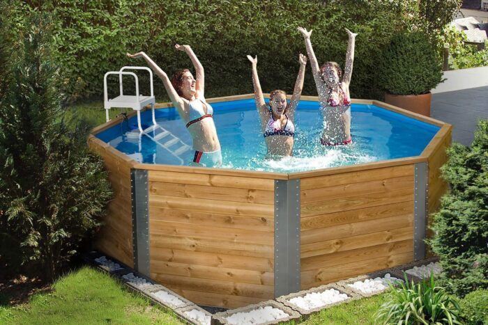 An eight-sided, wooden swimming pool in a garden. The swimming pool is filled with water and has a white ladder as an entrance. Three children are standing in the swimming pool and having fun.