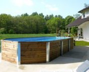 Basic solid wood pool from Weka. Large, angular swimming pool in natural wooden look. Stands in front of a large, white house.
