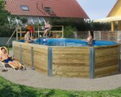 Qualitäts Swimmingpool von Weka. Grosser Pool im Garten mit einer Holztreppe. Pool steht vor einem grossen Haus mit roten Ziegelsteinen. Kinder spielen im Pool.