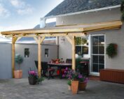 Naturbelassene Terrassenüberdachung mit drei verschiedenen Balken. Die Überdachung lehnt an einem weissen Haus an. Unter der Überdachung stehen dunkle Stühle und ein dunkler Tisch.