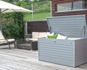 Offen stehende Gartenbox in silber mit verschiedenen Kissen verstaut. Die Gartenbox steht auf einer Terrasse mit Holzboden und Polyrattan Gartenlounge.