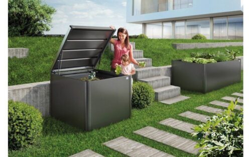 a dark grey composter in a garden, standing next to some steps and a woman holding the lid
