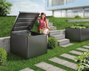 Ein dunkelgrauer Kompost in einem Garten vor einer modernen Villa mit Ganzglasfenster. Eine Frau hält den Deckel des Komposters offen und ein kleines Mädchen wirft Salatblätter in den Komposter hinein.