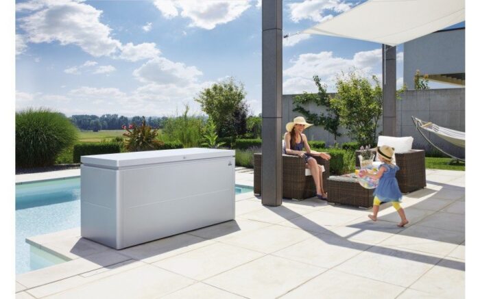Die biohort Loungebox. Abgebildet ist eine graue Metallbox die vor einem Pool steht auf einer Terrasse. Neben der Box ist eine Gartenlounge abgebildet mit einer Frau die auf einem Stuhl sitzt und ein Kind das vor ihr am spielen ist.