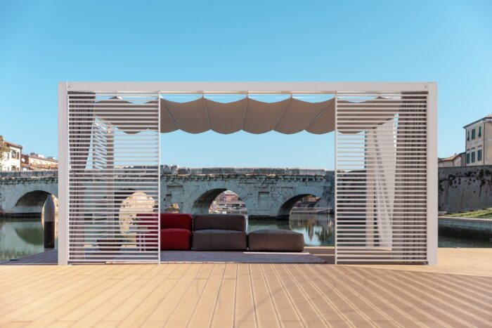 Unosider Pavillon Mood YPE mit zusammengebundenen Vorhängen und Dach. Der Pavillon steht auf einer Holzterrasse vor einer Brücke eines Flusses.