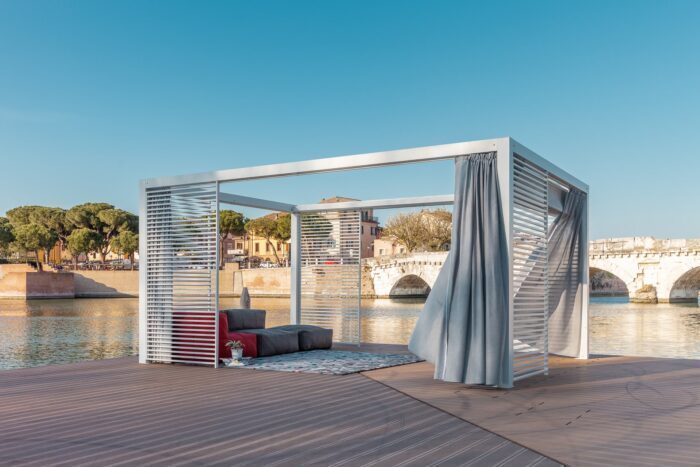 Unosider Mood YPE Pavillon mit grauen Vorhängen und geöffnetem Dach vor einem grossen Pool.