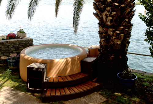 Ein brauner runder Softub Sportster mit Treppeneinstieg unter einer Palme vor einem See.