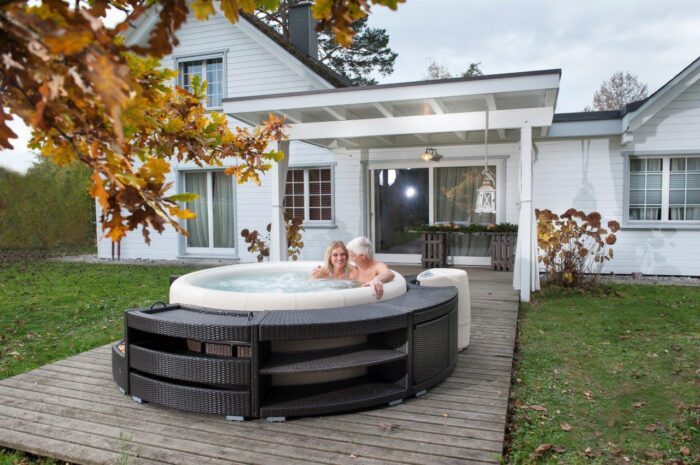 Resort soft tub whirlpool in off-white with black polyrattan edging. The Resort soft tub whirlpool stands on a wooden terrace in front of a large white house.